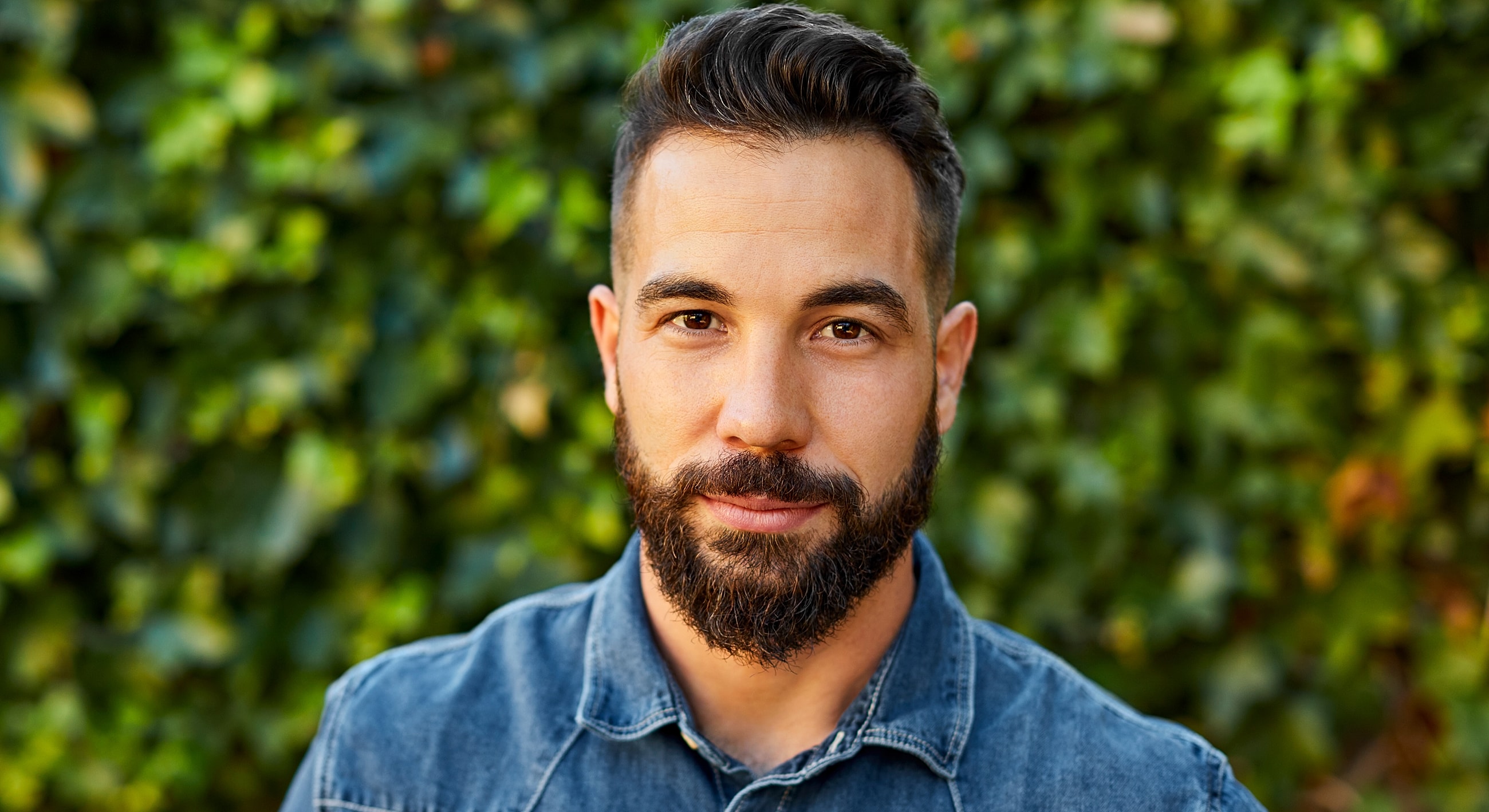Man with beard in denim shirt outdoors.